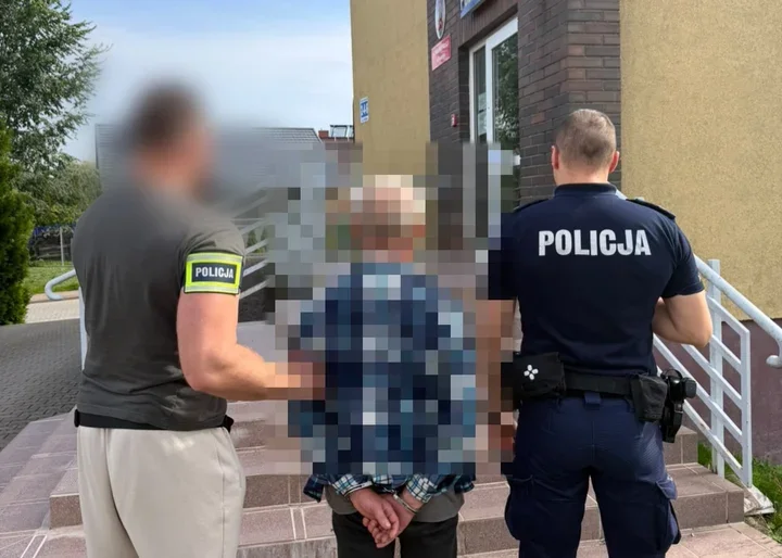 Dwóch policjantów prowadzi skutego mężczyznę przed budynek.