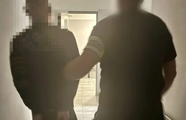 Dwie osoby w czarnych koszulkach stoją w wąskim korytarzu, w tle widoczne otwarte drzwi i podłoga z płytek.