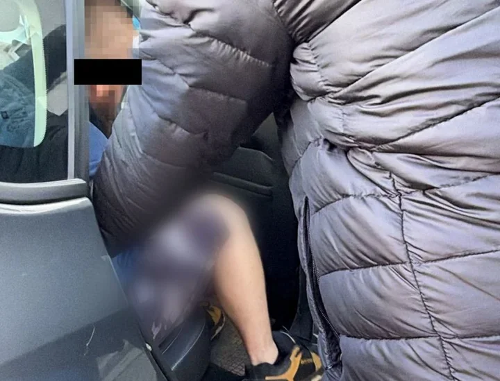 Osoba w szarej pikowanej kurtce wchodzi lub wysiada z pojazdu, przy otwartych drzwiach siedzi druga osoba.