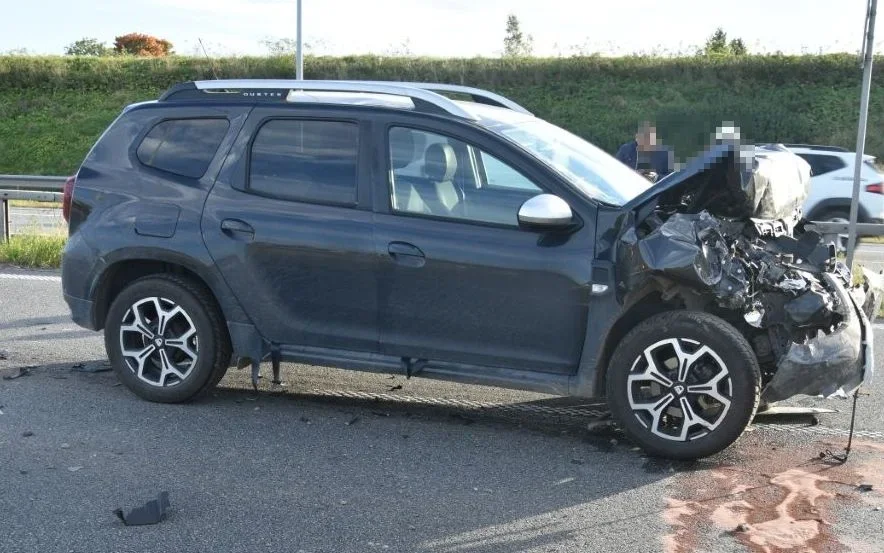 Uszkodzony po zderzeniu ciemny SUV Dacia Duster stoi na drodze, z widocznym zniszczeniem przedniej części pojazdu.