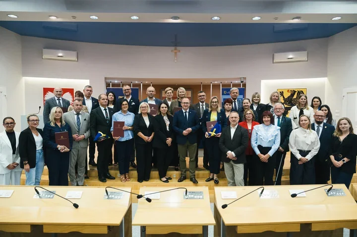 Grupa osób pozuje do pamiątkowego zdjęcia w sali konferencyjnej z drewnianymi stołami i mikrofonami, niektórzy uczestnicy trzymają teczki i dokumenty.