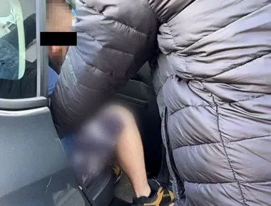 Osoba w szarej pikowanej kurtce wchodzi lub wysiada z pojazdu, przy otwartych drzwiach siedzi druga osoba.