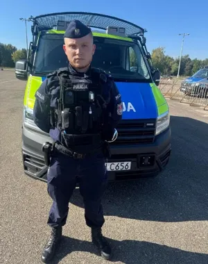 Funkcjonariusz policji w mundurze stoi przed policyjnym furgonetką na otwartym parkingu w słoneczny dzień.