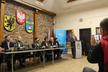 Mężczyzna w garniturze przemawia z mównicy podczas konferencji w sali z ceglanym sufitem, przy stole siedzi sześciu mężczyzn, a mężczyzna z prawej strony robi z