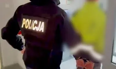 Funkcjonariusz policji w czarnej kurtce z napisem POLICJA idzie korytarzem obok rozmazanej postaci w żółtej koszulce.