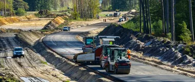 Robotnicy i maszyny budowlane układają asfalt na odcinku drogi w lesie, przygotowując nową dwujezdniową trasę.