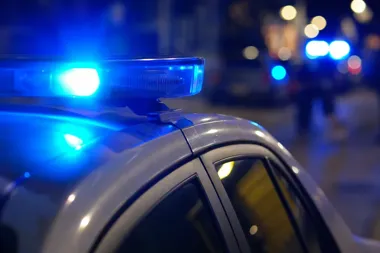 Nocna ulica z policyjnym radiowozem z włączonym niebieskim światłem na dachu, w tle rozmyte światła innych pojazdów.