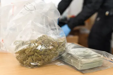 Woreczek strunowy z suszem roślinnym i torebka z banknotami leżą na drewnianym stole, w tle sylwetka osoby w ciemnym mundurze z niebieską rękawiczką.