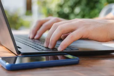 Dłonie osoby piszącej na klawiaturze laptopa, obok leży smartfon na drewnianym stole.