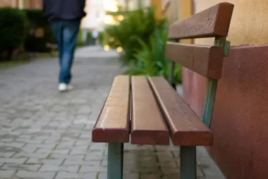 Drewniana ławka przy budynku na brukowanej alejce, w tle odchodząca osoba w dżinsach.
