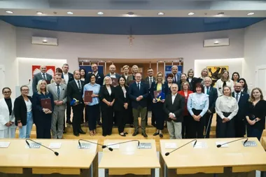 Grupa osób pozuje do pamiątkowego zdjęcia w sali konferencyjnej z drewnianymi stołami i mikrofonami, niektórzy uczestnicy trzymają teczki i dokumenty.